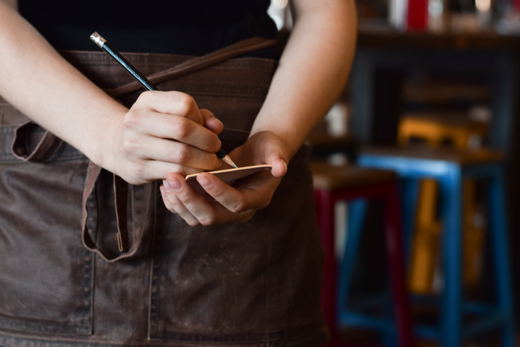 Tarjoilija kirjoittaa tilausta muistilapulle kynällä, yllään ruskea esiliina ja taustalla ravintolasali epätarkkana.