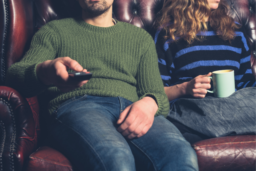 Kaksi aikuista istuu nahkasohvalla, toisella on kädessään kaukosäädin ja toisella muki