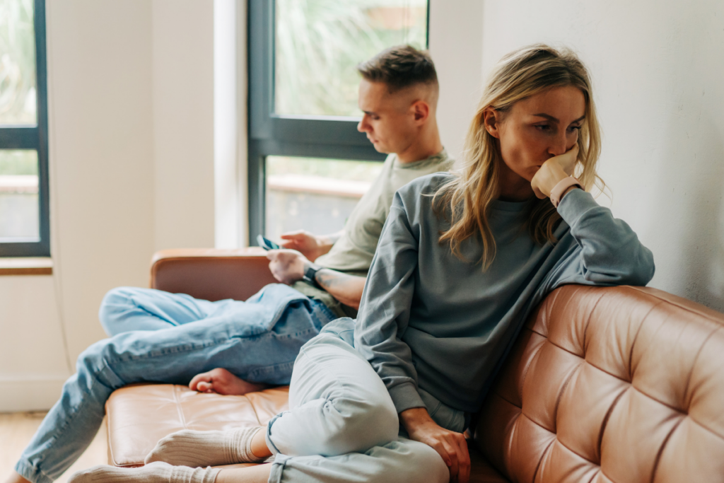 Kaksi ihmistä istuu sohvalla olohuoneessa, toinen nojaa sohvan selkänojaan ja toinen istuu taaempana katsoen puhelinta