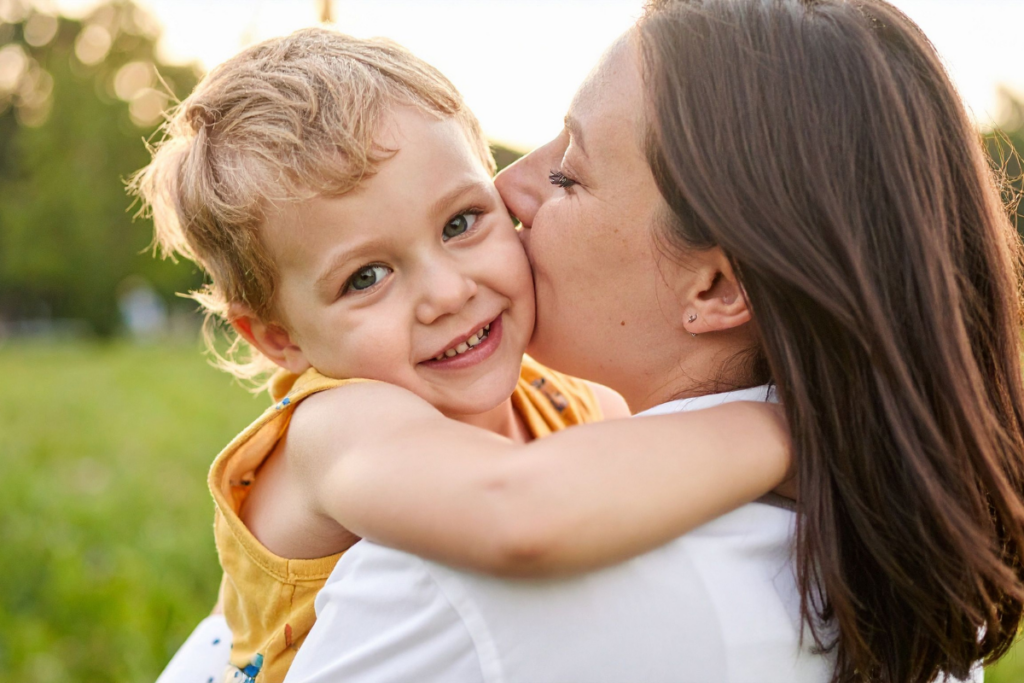 Vanhempi suutelee pientä lasta poskelle ulkona puistossa, lapsi hymyilee suoraan kameraan vanhemman sylissä.