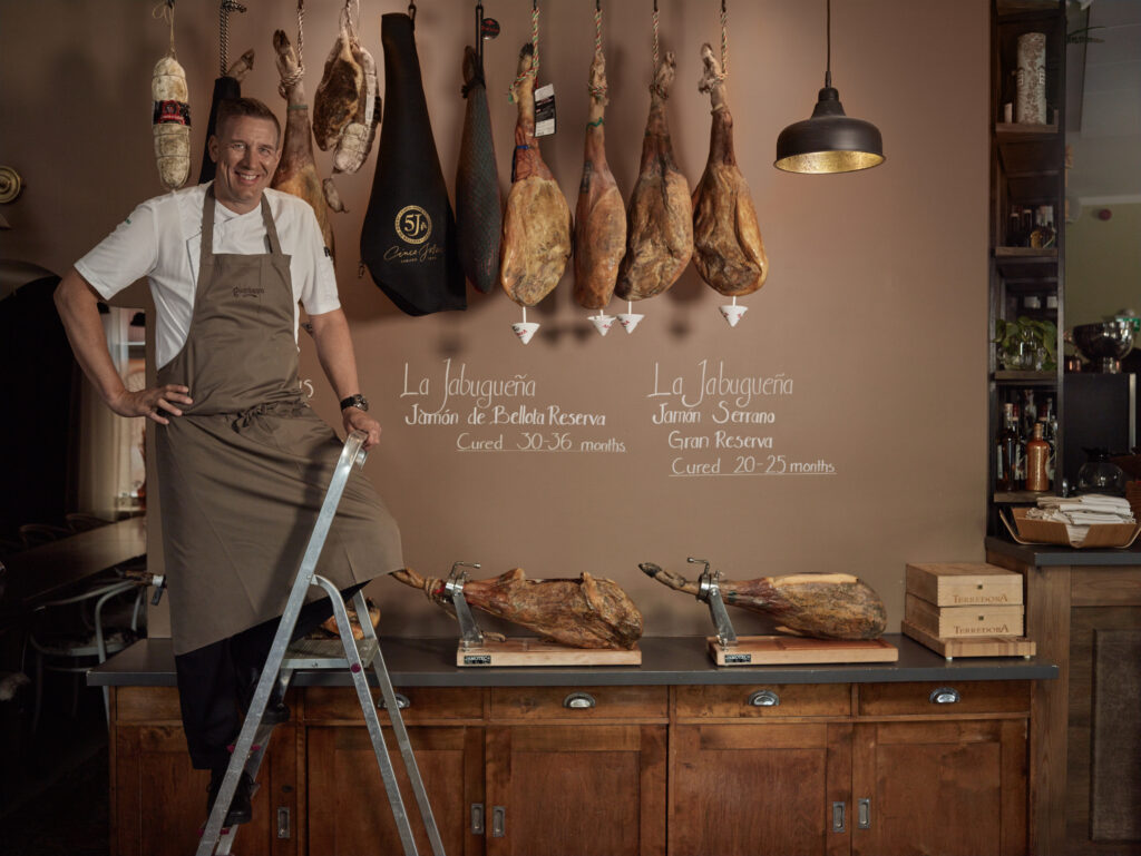 Kokki seisoo tikkailla ravintolassa, jossa seinällä roikkuu useita ilmakuivattuja kinkkuja.