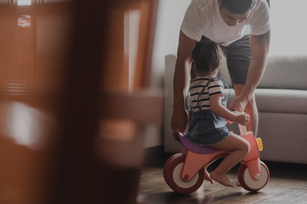 Isä auttaa pientä lasta ajamaan potkupyörällä kotona.