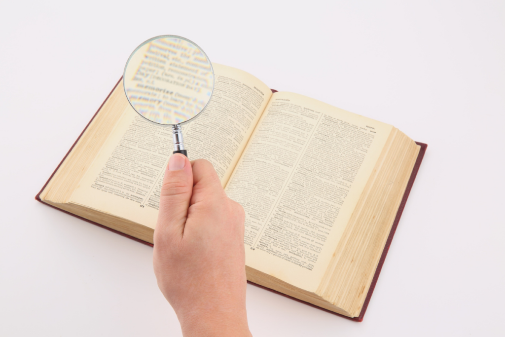 Käsi pitelee suurennuslasia avoimen kirjan päällä ja tarkastelee sivun tekstiä.