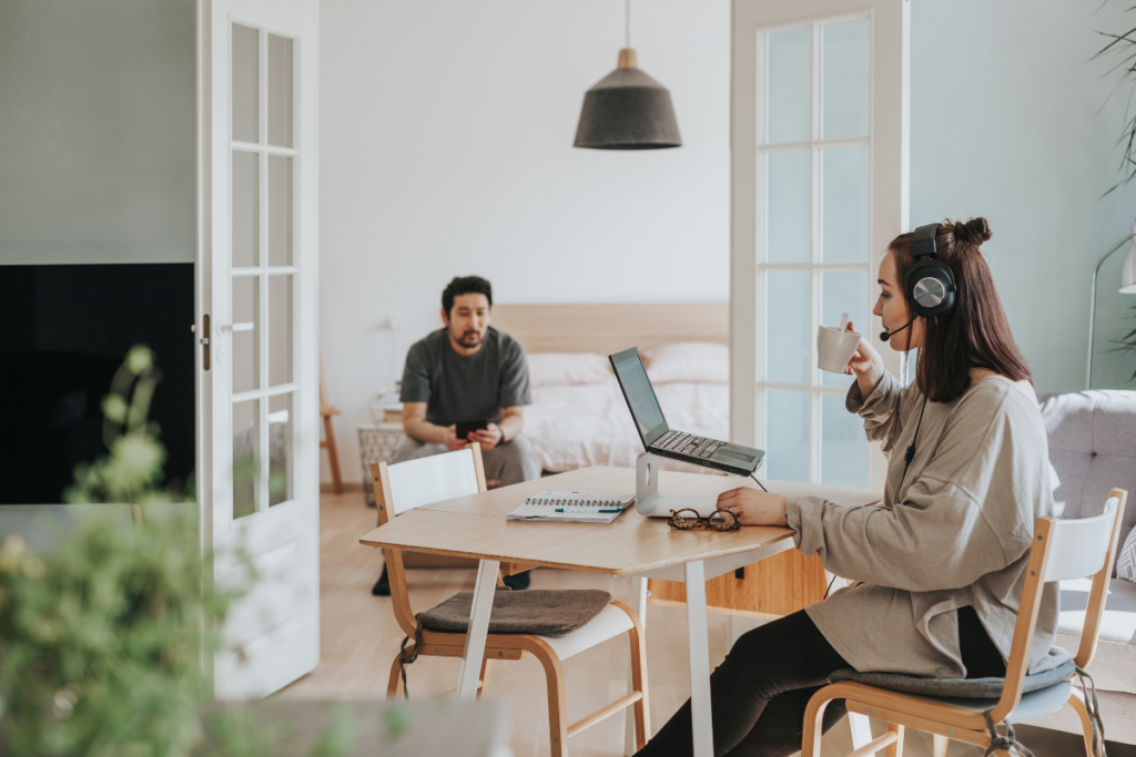 Nainen työskentelee läppärillä kuulokkeet päässä kotona, mies istuu taustalla sohvalla.