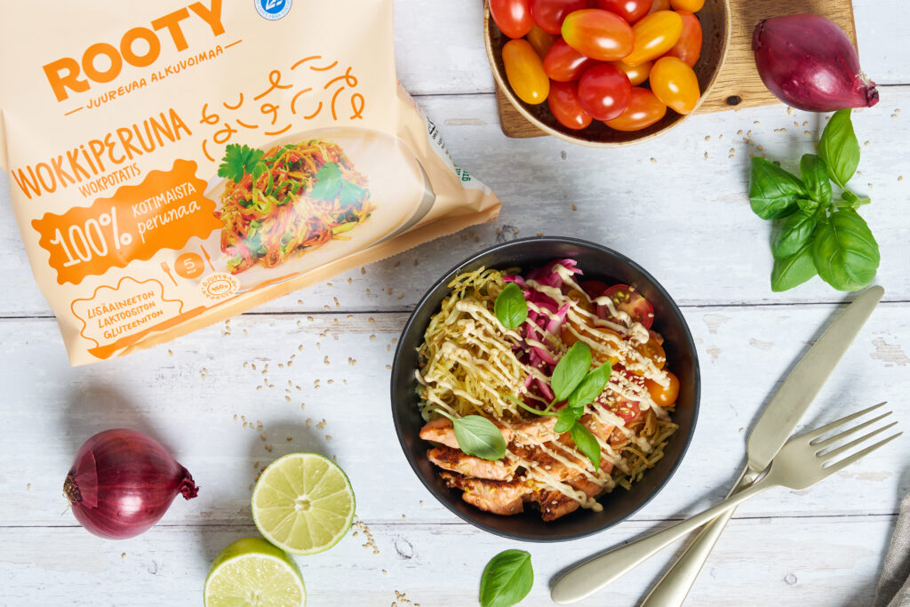 Rooty Wokkiperuna -pakkaus ja valmis wokkiperunabowl, jossa kanaa, kasviksia, yrttejä ja kastiketta; ympärillä tomaatteja, punasipulia, limeä ja ruokailuvälineet vaalealla pöydällä.