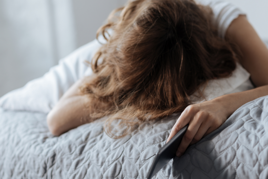 Väsynyt nainen makaa sängyssä ja pitää puhelinta kädessään.