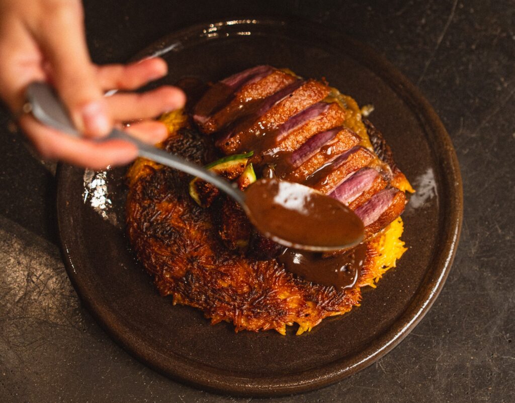 Viipaloitu ankka-annos rapean perunapohjan päällä, jonka päälle lusikoidaan tummaa kastiketta; annos kuvattu lähietäisyydeltä ravintolassa.