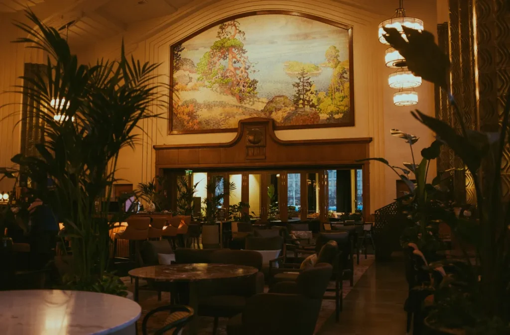 Taulu-ravintolan ravintolasali, jossa on runsaasti kasveja, pyöreitä pöytiä ja pehmeää valaistusta; taustalla suuri maisemataulu seinällä.