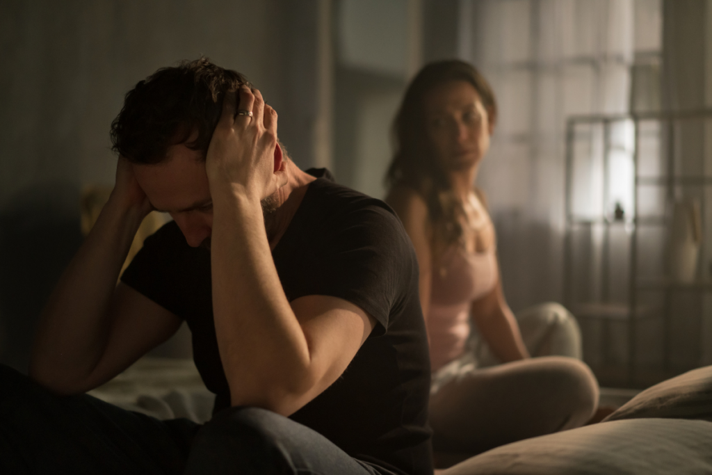 Mies istuu sängyn reunalla pää käsissään, nainen istuu taustalla vakavana – riita tai jännittynyt tilanne parisuhteessa.